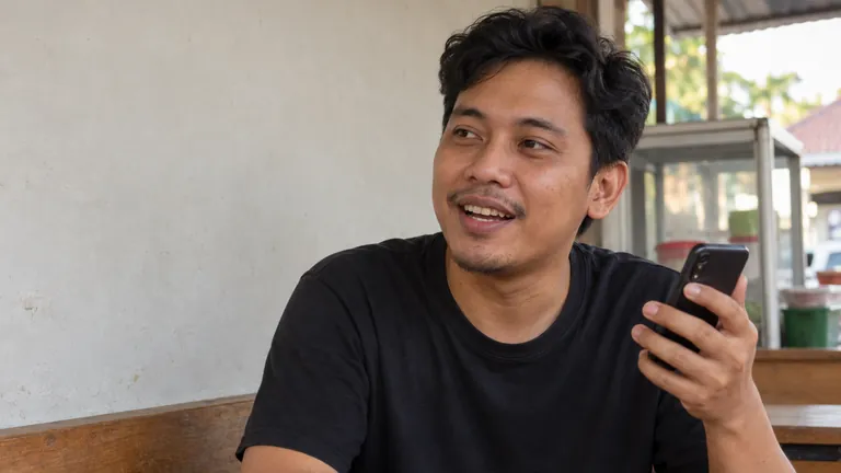 Pria tersenyum santai memegang HP di bangku warung