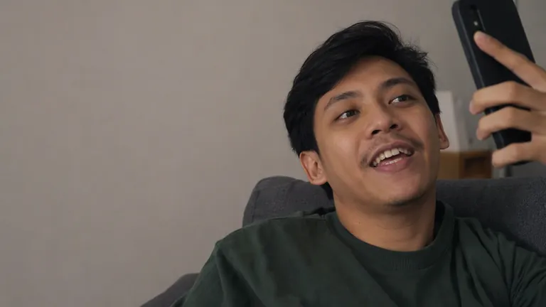 Pria rebahan di sofa tersenyum melihat HP